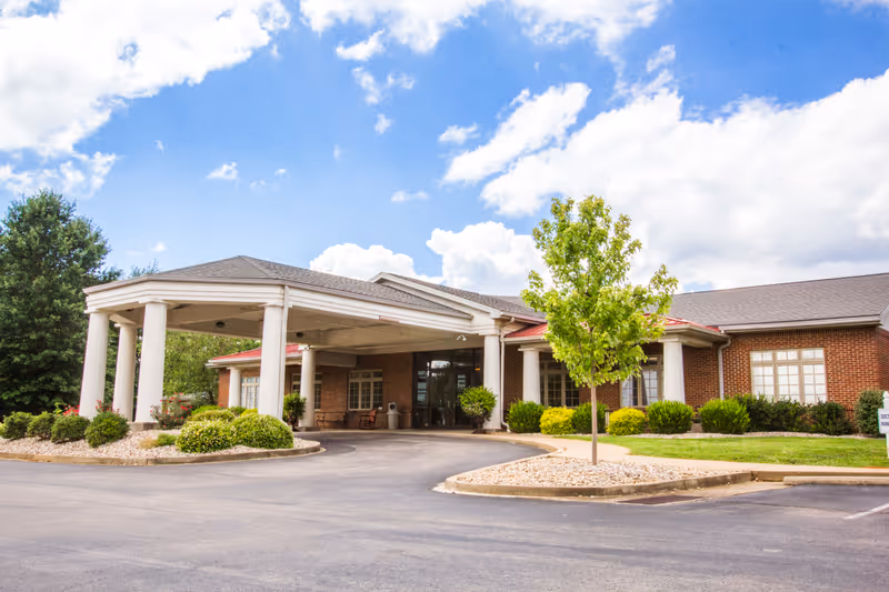 Exterior view of a single-story brick building with a covered entrance supported by white columns. The building is surrounded by neatly trimmed bushes, a small tree, and a paved driveway under a partly cloudy blue sky.