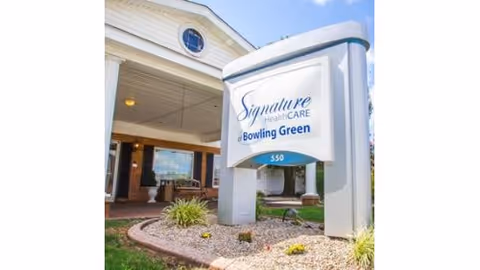 Entrance area of Signature HealthCARE of Bowling Green with a large white sign displaying the facility name and address number 550, set in a landscaped area with rocks and plants, and part of the building with columns and a covered porch visible in the background under a blue sky.