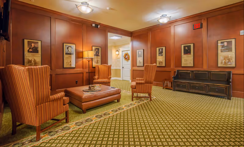 A cozy lounge with striped wingback chairs, a leather ottoman, wood-paneled walls and framed portraits.