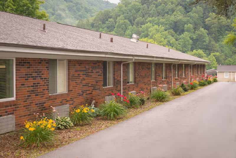 Long side view of a single-story brick building with multiple windows, a paved driveway, and flower beds with yellow and red flowers along the base. The building is surrounded by lush green trees and hills in the background.