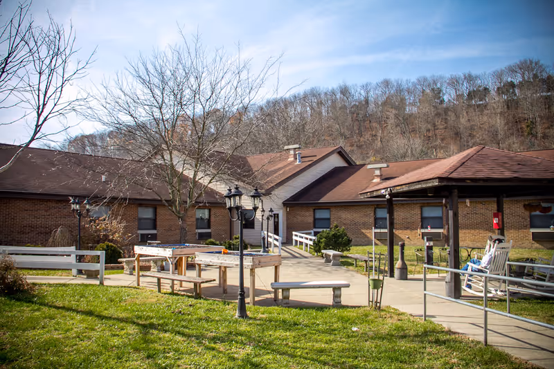 Brick one-story senior living facility courtyard with picnic tables, benches, lamp posts, a covered patio and a grassy lawn under leafless trees.
