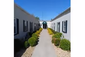 Sunlit walkway between white buildings, bordered by trimmed shrubs and gravel landscaping.