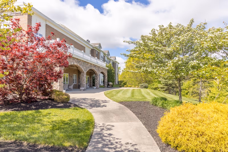 A bright outdoor scene showing a paved walkway curving alongside a senior living community building with stone arches. The area is landscaped with green grass, a red-leafed tree, a flowering tree, and yellow shrubs under a partly cloudy blue sky.