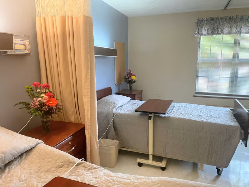 A nursing facility room with two hospital beds separated by a beige privacy curtain. Each bed has a light-colored quilted bedspread, and there are bedside tables with flower vases. A window with blinds and a floral valance lets in natural light.