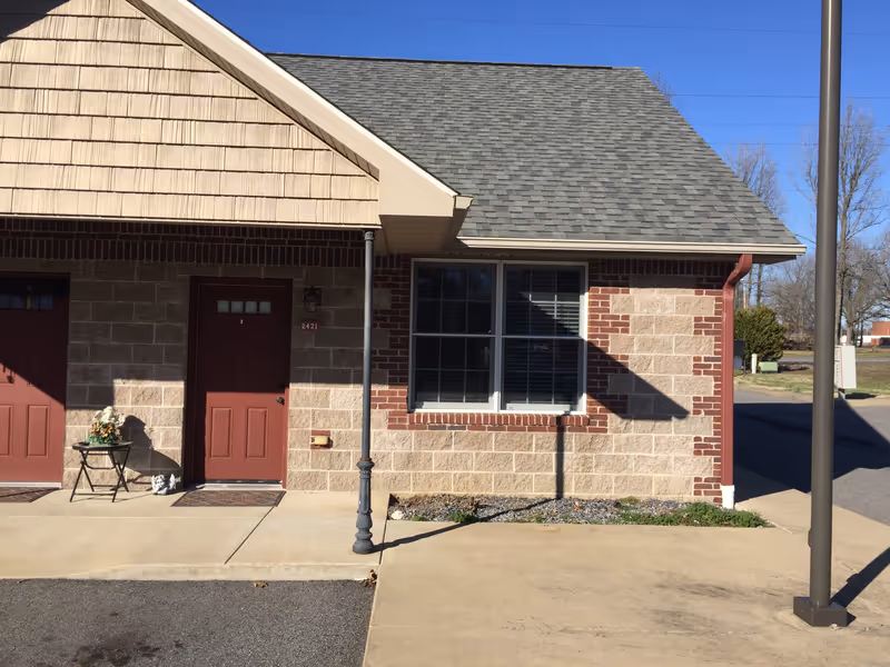 Exterior view of a building with beige brick and red brick accents, two red doors, a window with closed blinds, a small table with a flower arrangement, and a street lamp post on the sidewalk.