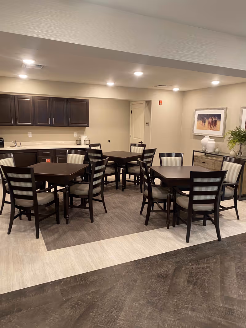 Well-lit dining area with several dark wood tables and striped cushioned chairs in a kitchenette/lounge space.