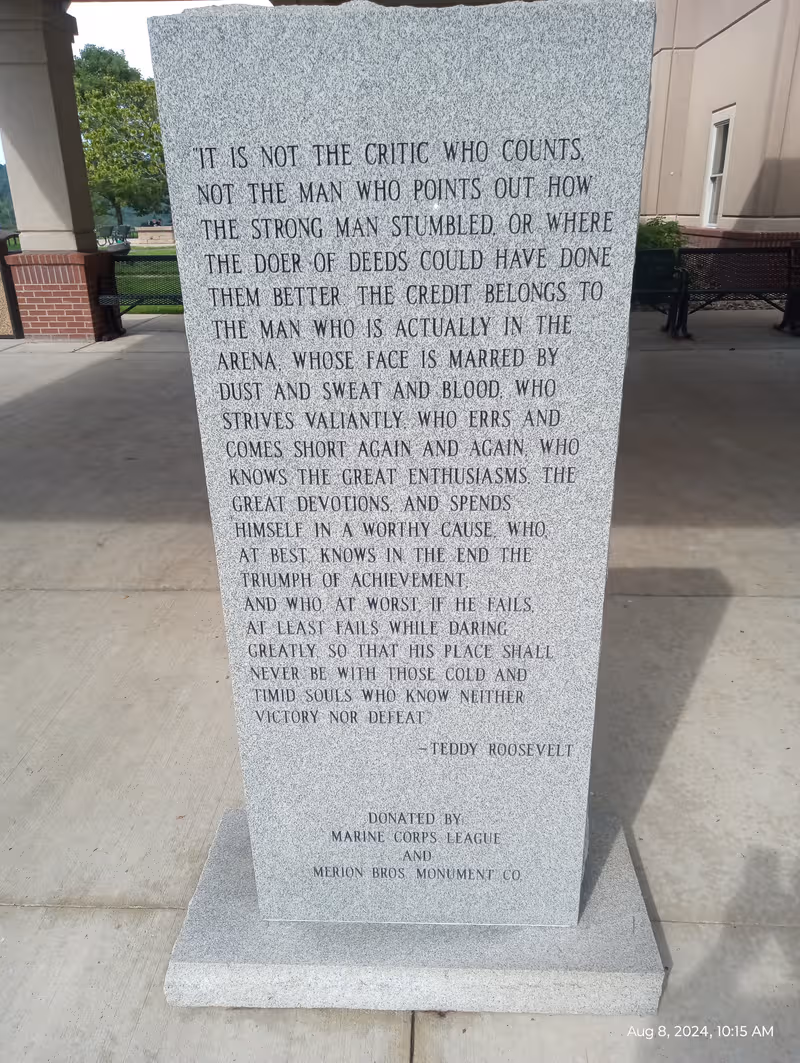 Tall granite memorial stone with Teddy Roosevelt quote set outside a building.