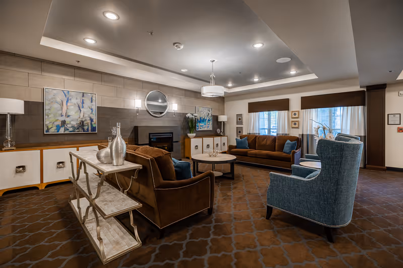 A cozy living room area in a senior living facility featuring two brown sofas with blue accent pillows, two blue upholstered armchairs, a round coffee table, and a console table with decorative silver vases. The room has a patterned brown carpet, a modern ceiling light fixture, wall-mounted lights, a round mirror above a fireplace, and abstract artwork on the walls. Large windows with brown valances and white curtains allow natural light into the space.