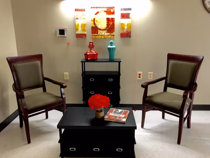 A small sitting area with two wooden armchairs with green cushions facing a black coffee table. On the coffee table is a small pot of red flowers and a book. Behind the table is a black cabinet with drawers, decorated with two ornamental vases, one red and one turquoise. Above the cabinet are three abstract art pieces in red, yellow, and white tones. The walls are beige and the floor is light-colored tile.