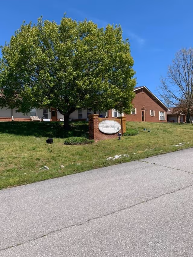 Exterior view of Twin Oaks Assisted Living facility showing a large green tree, a brick sign with the facility name, a grassy lawn, and a paved driveway under a clear blue sky.