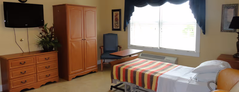 A senior living bedroom with a single bed covered by a striped blanket, wooden dresser and wardrobe, wall-mounted TV, armchair and a bright window.