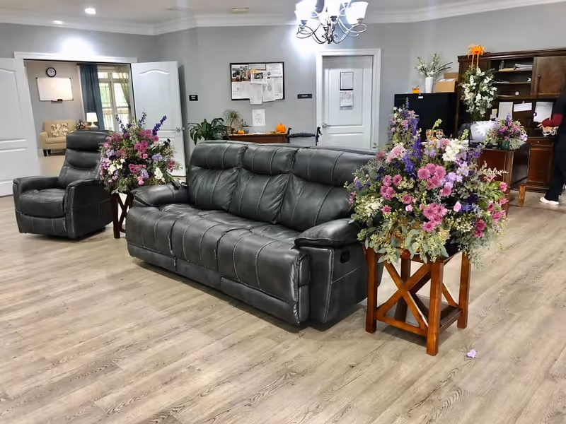 A senior living facility common area with a gray leather sofa and matching armchair on a wooden floor. Two wooden stands hold large floral arrangements with purple, pink, and white flowers. In the background, there is a wooden desk with a person standing nearby, a bulletin board, and a doorway leading to another room with a beige couch and a clock on the wall.