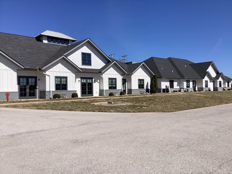 Exterior view of a single-story senior living facility building with white siding, multiple windows, and a dark roof under a clear blue sky. There is a paved driveway and a grassy area in front of the building.