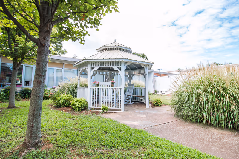 White wooden gazebo with rocking chairs in a landscaped courtyard with trees and ornamental grasses outside a care building.