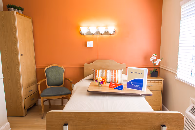 A single bed with a wooden headboard and white bedding is placed against an orange accent wall. On the bed is a wooden tray holding a welcome book, a small flower vase, and some informational cards. To the left of the bed is a wooden wardrobe and a green cushioned chair with wooden arms. To the right is a wooden nightstand with a white orchid plant. A window with white blinds allows natural light into the room. Above the bed is a wall-mounted light fixture with five bulbs.