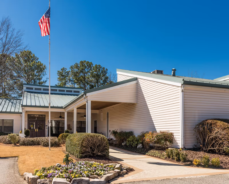 Exterior view of Louisville East Post Acute Care facility showing a single-story building with white siding and a green metal roof. There is a covered entrance with columns, a flagpole with an American flag, landscaped bushes, and a flower bed in the foreground under a clear blue sky.