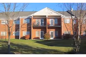 Two-story red-brick senior living building front with second-floor balconies, ground-level patios, and a grassy lawn with small trees.