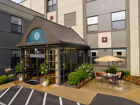 Entrance to a building with a glass vestibule and a brown metal roof, surrounded by green bushes and flowers. There is a small outdoor seating area with a table, chairs, and a beige umbrella on a concrete patio. The building exterior is beige and gray with multiple windows.