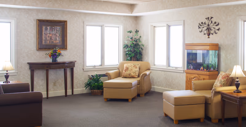 A cozy living room area with beige armchairs and matching ottomans, a brown sofa, two side tables with lamps, a wooden console table with a vase of flowers, two potted plants, a fish tank on a wooden stand, and framed artwork on the wall. The room has three windows letting in natural light and neutral-colored walls and carpet.