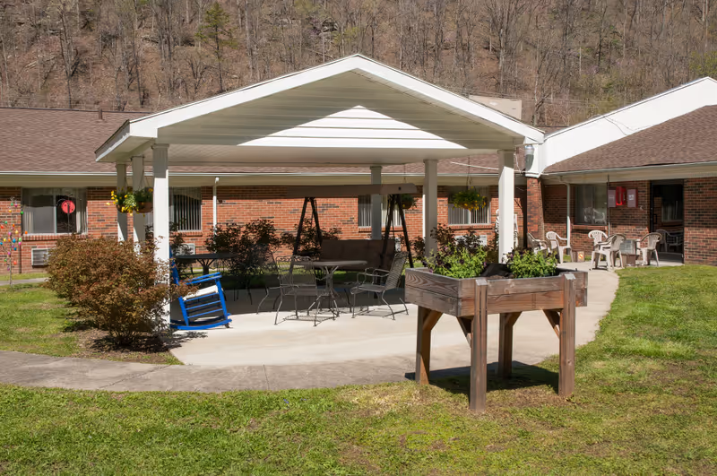 Outdoor covered patio with metal chairs and tables, brick building behind, wooden planter in foreground.