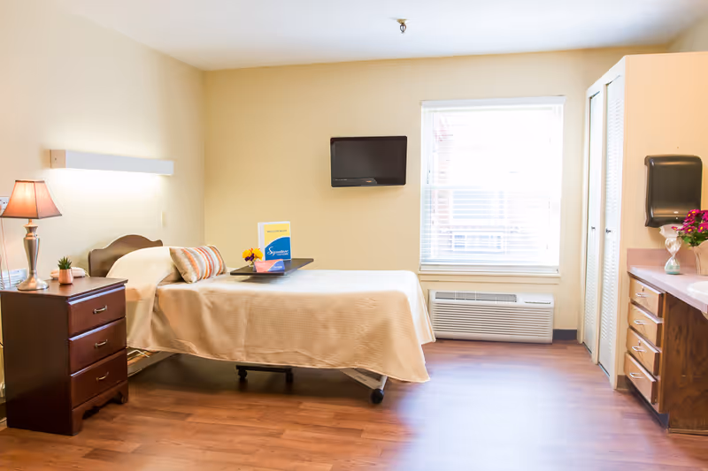A clean and bright bedroom in a healthcare facility featuring a single bed with a beige bedspread, a bedside table with a lamp and small plant, a wall-mounted TV, a window with blinds, and a countertop with drawers and a paper towel dispenser.