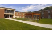 Two-story brick senior living building surrounding a grassy courtyard with a pergola and paved walkway under a partly cloudy sky.