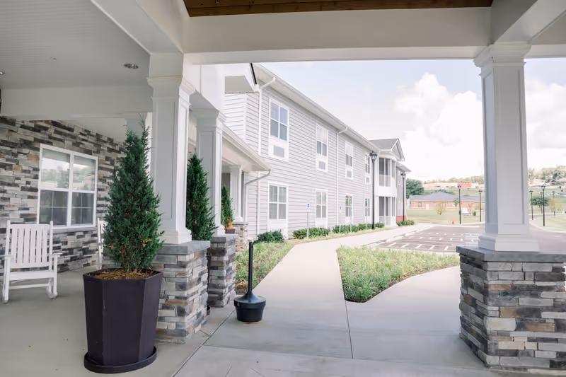 Covered front entrance and walkway of a senior living building with columns, planters, a rocking chair, and a parking area.