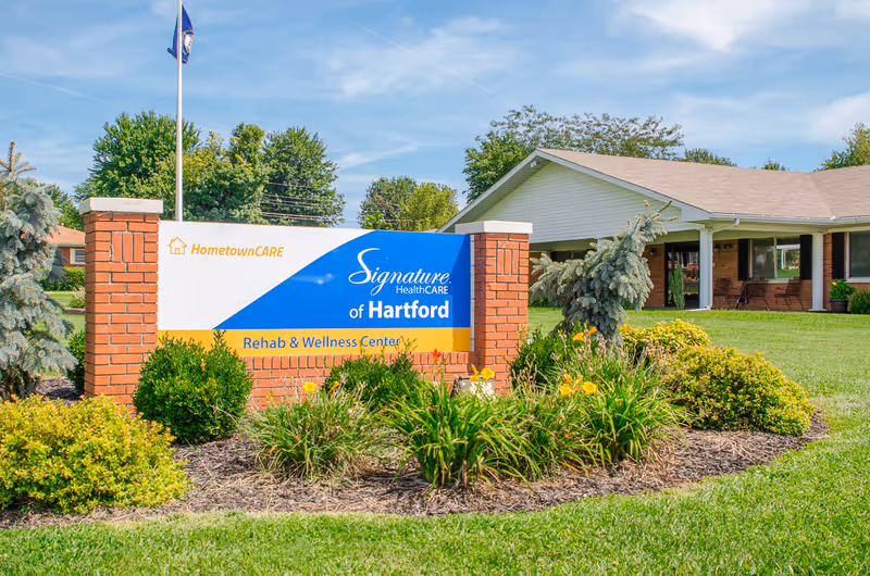 Outdoor view of the entrance sign for Signature HealthCARE of Hartford Rehab & Wellness Center, surrounded by green bushes and yellow flowers, with a building and trees in the background under a blue sky.