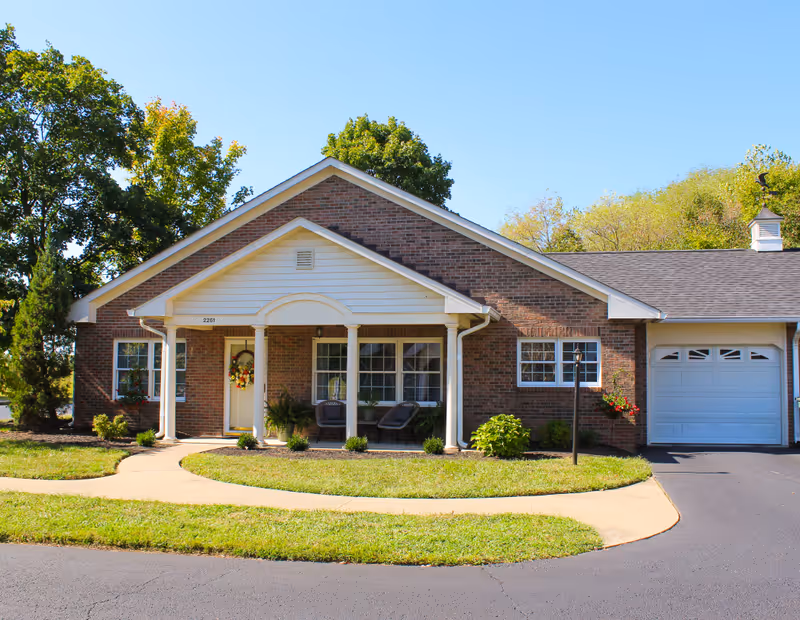 Single-story brick house with a white front porch supported by columns, two chairs on the porch, a front door decorated with a wreath, a garage on the right side, and a curved walkway leading to the entrance. The house is surrounded by green grass, small shrubs, and trees under a clear blue sky.