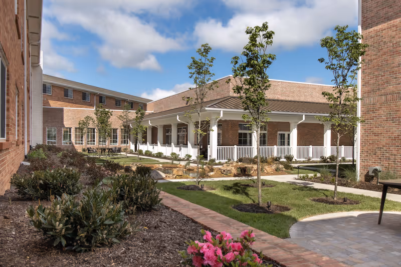 Outdoor courtyard area of a senior living facility with brick buildings surrounding a landscaped garden. The garden features small trees, shrubs, a paved walkway, and a covered patio with white railings. The sky is partly cloudy with blue patches visible.