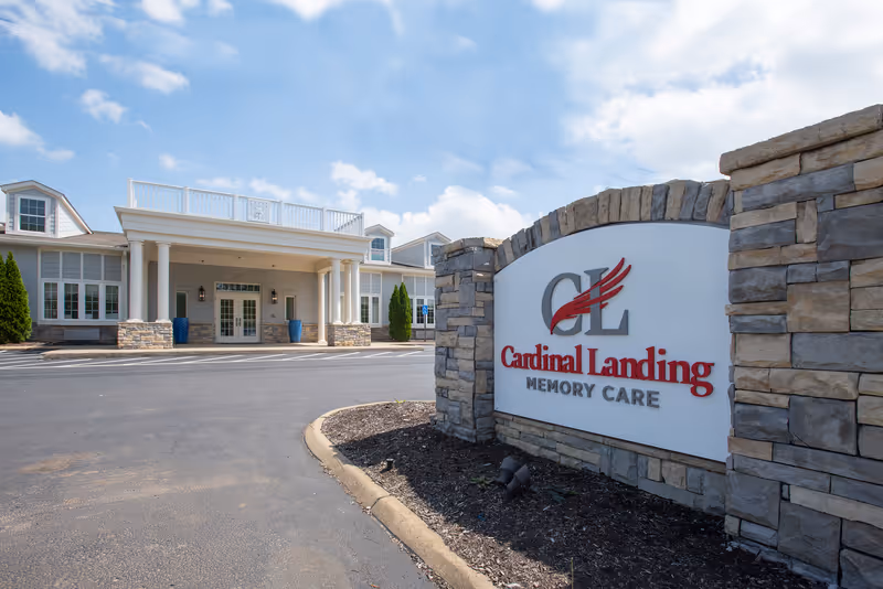 Exterior view of Cardinal Landing Memory Care facility showing the main entrance with white columns and a stone sign with the facility's name and logo in the foreground under a partly cloudy sky.