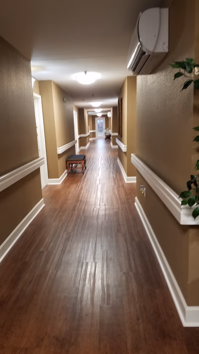 Long, well-lit hallway in a senior living facility with brown walls, wood floor, and bench.
