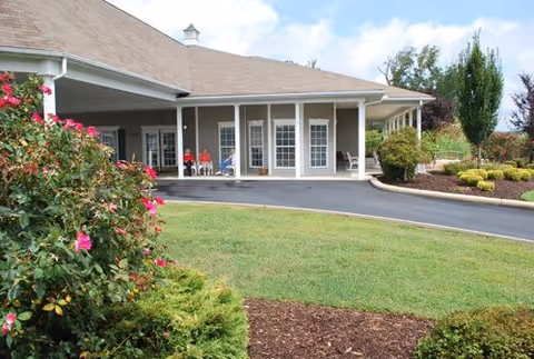 Exterior view of a single-story building with a covered entrance and large windows. There are three people sitting on chairs near the entrance. The surrounding area includes a paved driveway, green lawn, bushes, and flowering plants under a partly cloudy sky.