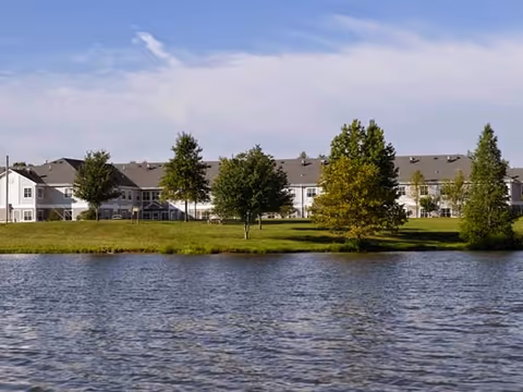 View of a large senior living facility building behind a grassy area with several trees, seen across a body of water under a partly cloudy sky.