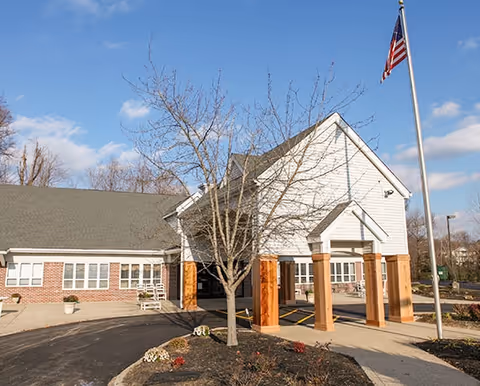 Exterior view of a senior living facility building with a covered entrance supported by wooden columns, a leafless tree in front, and an American flag on a flagpole to the right under a partly cloudy sky.
