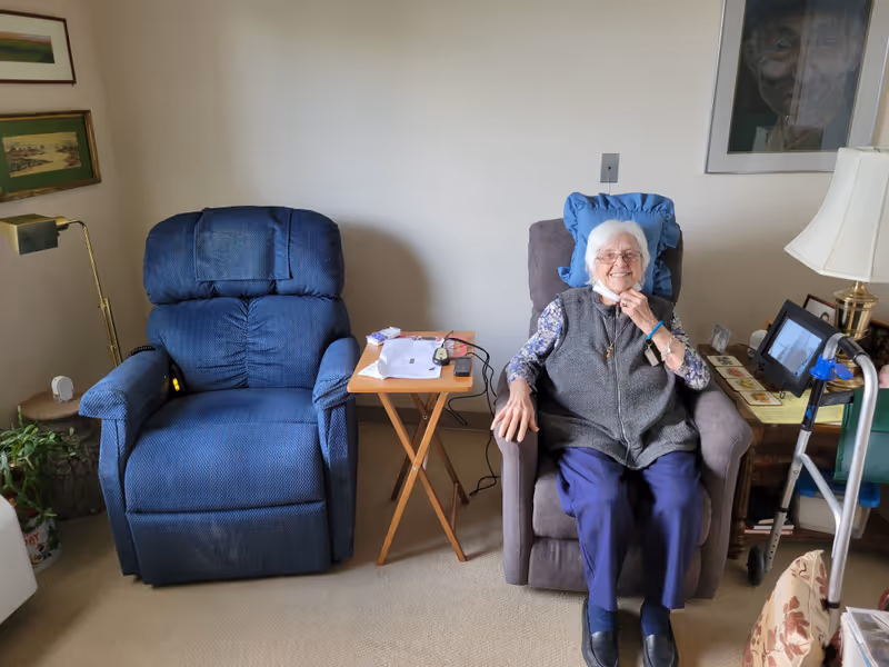 An elderly woman sits in a cushioned recliner in a living room with a side table.