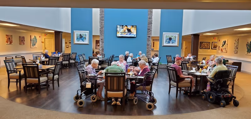 Seniors seated at multiple tables eating and socializing in a large communal dining room with a TV on a blue accent wall.