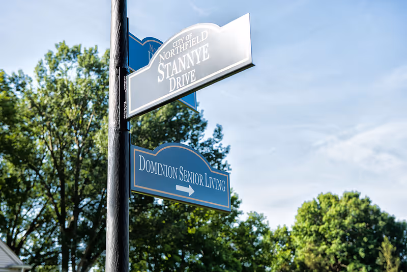 A pole-mounted street sign points toward 'Dominion Senior Living' and 'Stannye Drive' with trees and a blue sky in the background.