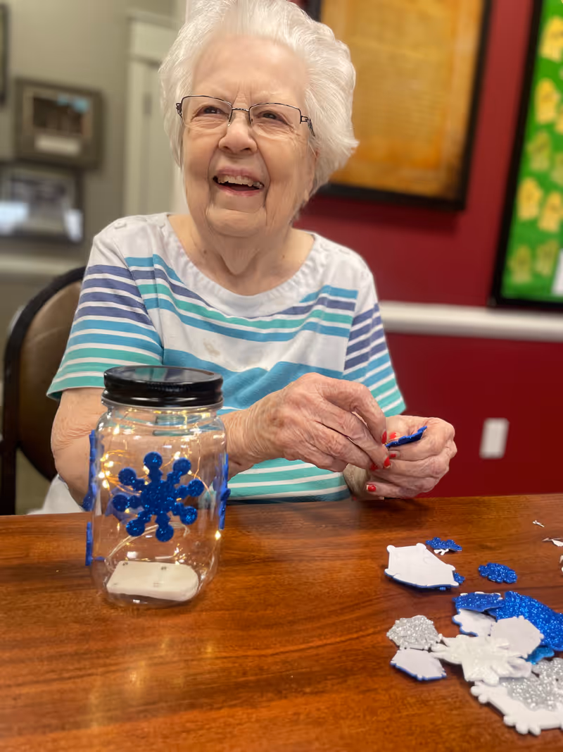 An elderly woman with white hair and glasses is sitting at a wooden table, smiling while working on a craft project involving blue and white snowflake decorations and a clear jar with a black lid. The background shows a red wall with framed artwork and a bulletin board.