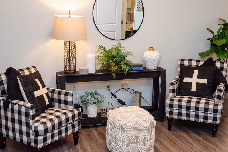 A cozy living room corner with two black and white checkered armchairs, each with a black pillow featuring a white cross design. Between the chairs is a black wooden console table with decorative items including a lamp, plants, vases, and books. A round mirror hangs on the wall above the table, and a textured white pouf sits on the wooden floor in front of the table.