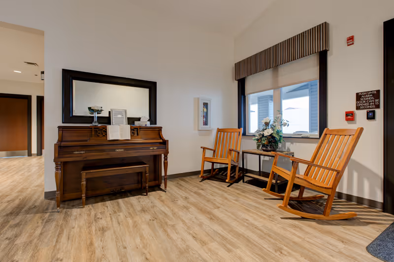 A cozy interior corner of a senior living facility featuring a wooden upright piano with sheet music and a vase on top, a large rectangular mirror hanging above it, two wooden rocking chairs, a small table with a floral arrangement, and a window with a striped valance. The floor is light wood, and there are safety signs and a fire alarm on the wall.