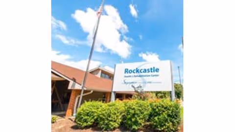 Exterior view of Rockcastle Health and Rehabilitation Center building with a sign displaying the facility's name in front, surrounded by green bushes and an American flag on a flagpole against a blue sky with clouds.