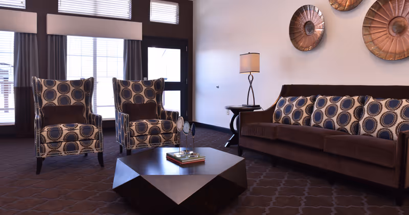 A cozy living room area with two patterned armchairs and a brown sofa adorned with matching patterned cushions. A modern geometric coffee table with books and decorative items sits in the center. A side table with a lamp is placed next to the sofa, and two large decorative metal wall hangings are mounted on the wall. Large windows with blinds and curtains allow natural light into the room.
