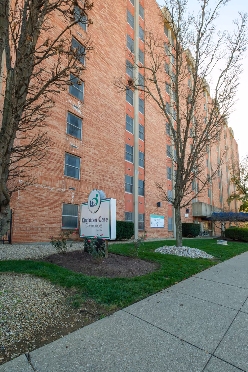 Exterior view of a multi-story brick building with several windows and leafless trees in front. A sign near the sidewalk reads 'Christian Care Communities'. The building has a concrete sidewalk and landscaped areas with grass and small bushes.