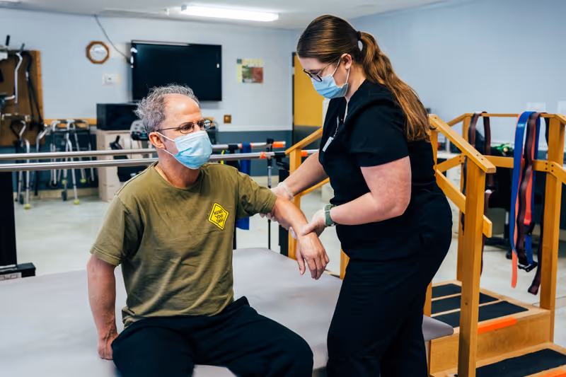 A healthcare professional wearing a mask and gloves assists an elderly man, also wearing a mask, with arm exercises in a physical therapy room equipped with parallel bars and exercise bands.