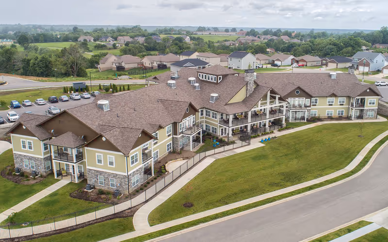 Aerial view of a large senior living facility building with multiple balconies, surrounded by a well-maintained lawn, sidewalks, and a parking lot. The building has a brown roof and beige siding with stone accents. Residential houses and greenery are visible in the background under a partly cloudy sky.