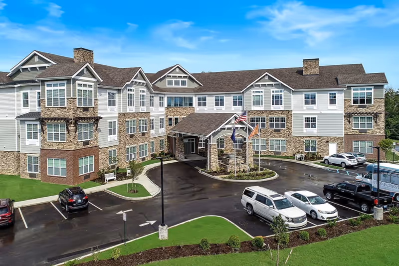 Three-story senior living building front with stone and siding facade, a covered entrance with flagpoles, and a parking lot with several cars.