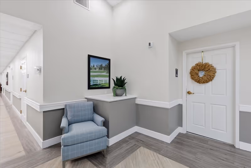 Senior living facility hallway with a blue armchair, plant, framed art, and a door with wreath.