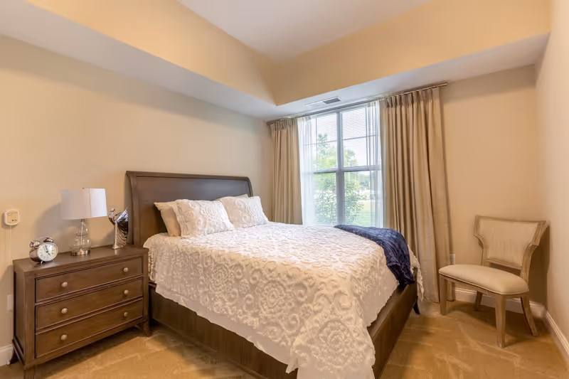 A bedroom with a wooden bed frame and a white textured bedspread, two pillows, a dark blue throw blanket, a wooden nightstand with a lamp, an alarm clock, and a decorative item. There is a window with beige curtains and a beige upholstered chair in the corner.