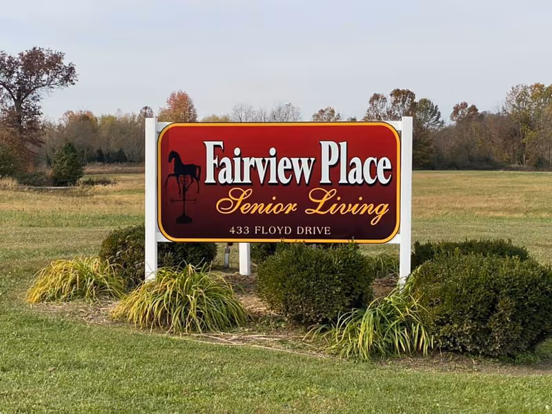 A large red and yellow sign for Fairview Place Senior Living at 433 Floyd Drive, set in a grassy area with bushes and trees in the background under a cloudy sky.
